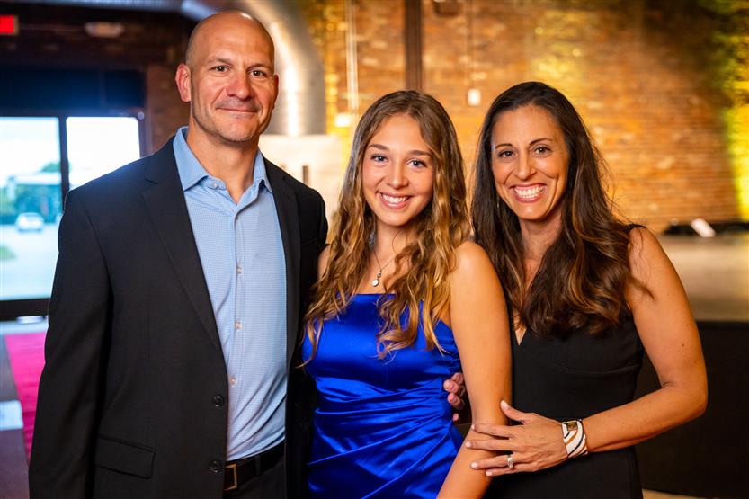 A man in a black blazer, a woman in a blue dress and a woman in a black dress smile